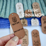 Hand holding Faux leather tag in color cinnamon or light brown, with the text Crochet by custom name and holes for attaching to crocheted or knitted items. And more tags with personalized texts and logos on the background.