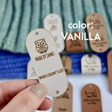 Hand holding Faux leather tag in color vanilla or off white, with the text Crochet by custom name and holes for attaching to crocheted or knitted items. And more tags with personalized texts and logos on the background.