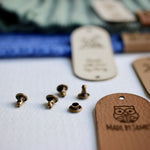 Close up of the rivets used to attach faux leather tags to handmade items, the rivets have 2 parts that are joined together to attach the tags. The color of the rivets for tags is bronze. Tags for crochets and knits with custom text and logos on the background.