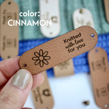 Hand holding Faux leather tag in color cinnamon or light brown, with the text Crochet by custom name and holes for attaching to crocheted or knitted items. And more tags with personalized texts and logos on the background.