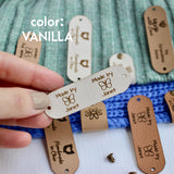 Hand holding Faux leather tag in color vanilla or off white, with the text Crochet by custom name and holes for attaching to crocheted or knitted items. And more tags with personalized texts and logos on the background.